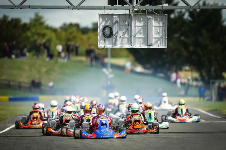 Pack of karts at the start beneath an LED screen hung from a trackside gantry