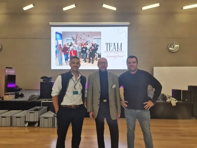 Trois hommes posent dans une salle de conférence devant un écran de projection affichant une présentation
