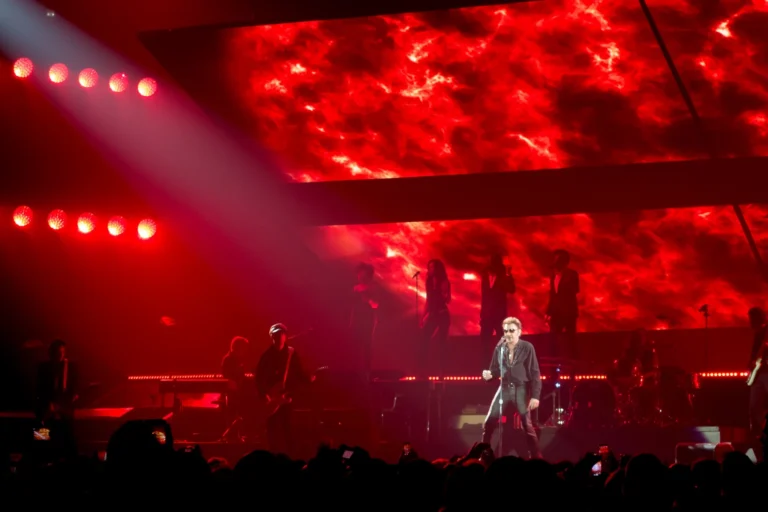 Singer centre stage with band and backing vocalists before red flame imagery on the LED screens