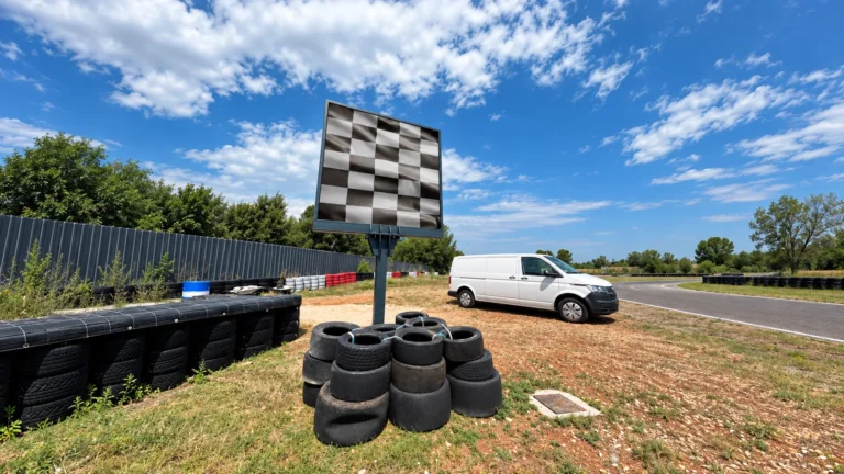 Square pole-mounted LED display showing a chequered flag beside the kart track under blue sky