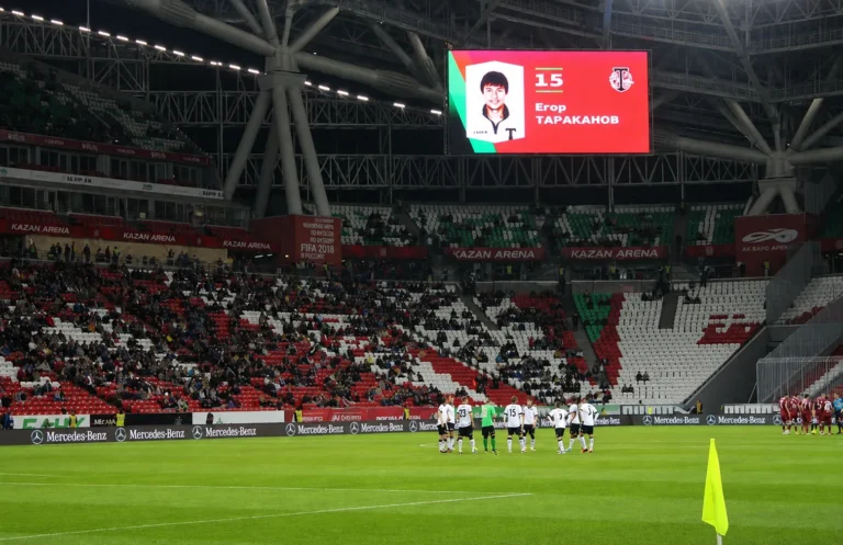 Giant LED screen hung under the roof showing a player, pitch and filled stands below
