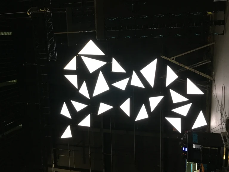Triangular LED panels lit white in the darkened venue, stage rig standing all around them