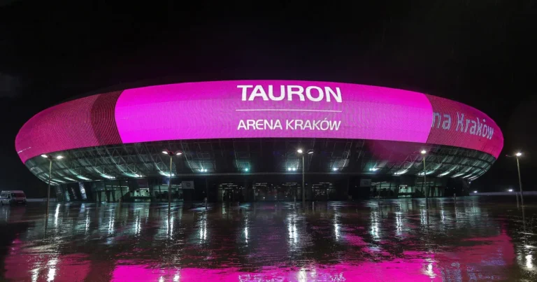 Façade LED de la Tauron Arena Kraków illuminée en rose la nuit, reflets sur le parvis mouillé