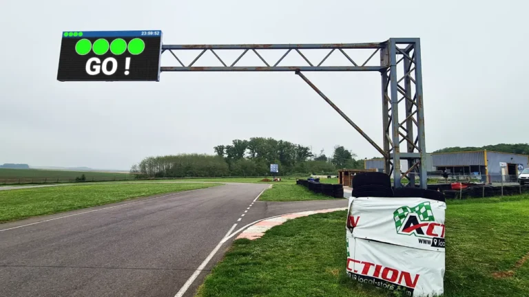 Écran LED monté sur une poutre métallique enjambant la piste de karting, sous un ciel couvert